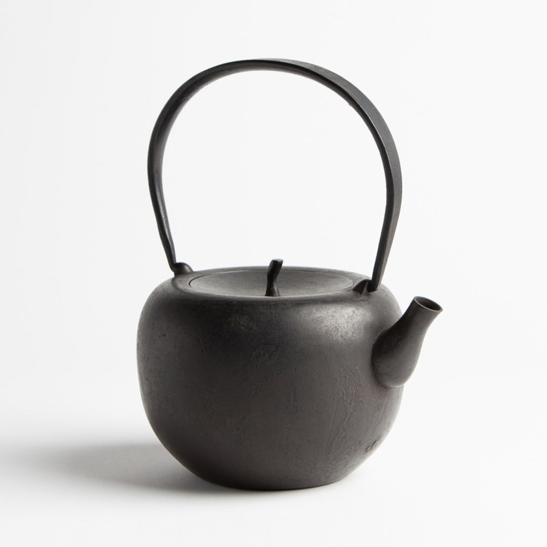 A black cast iron kettle on a white backround. The kettle is round with a flattish top and a lid a bit like the top of an apple. The kettle has a long handle for holding on top.
