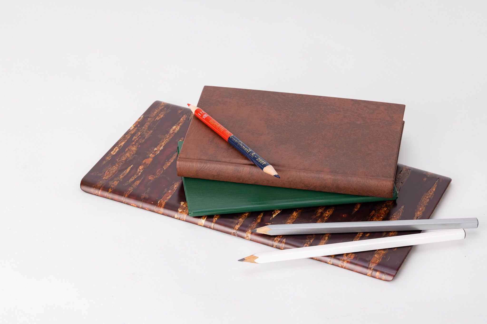 A rectangular wooden plate made of shiny red and orange cherry bark. On the plate are a white and silver pencil, a thin, green leather-bound notebook, a thicker brown leather-bound notebook, and on on top of the brown book, a pencil crayon with one red end and one navy blue end.