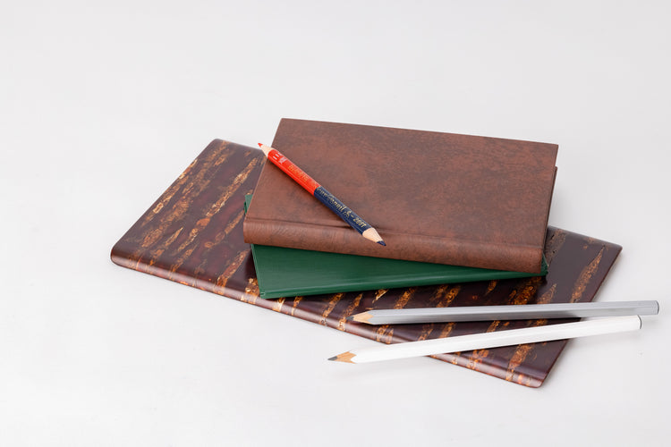 A rectangular wooden plate made of shiny red and orange cherry bark.  On the plate are a white and silver pencil, a thin, green leather-bound notebook, a thicker brown leather-bound notebook, and on on top of the brown book, a pencil crayon with one red end and one navy blue end.