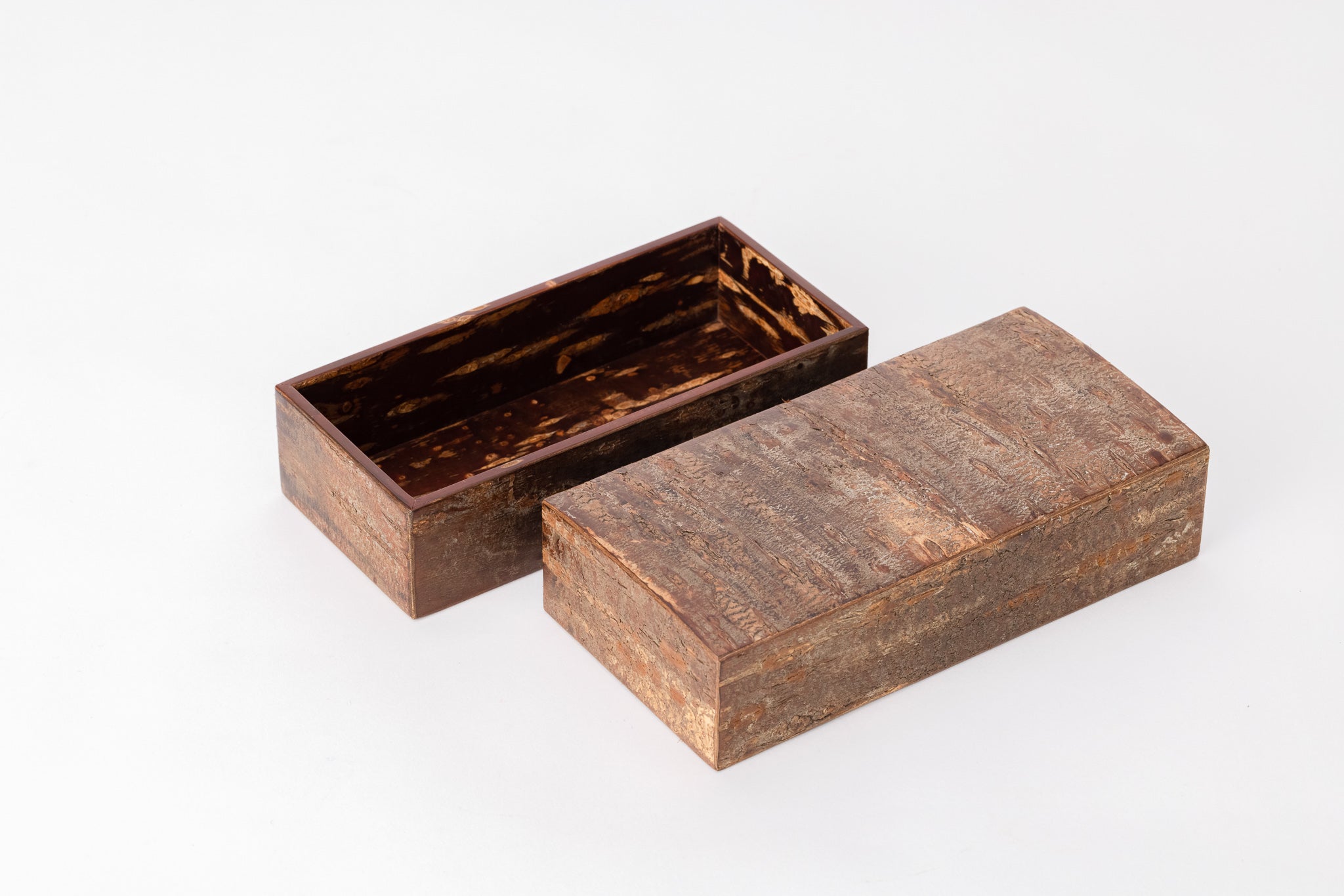 A rectangular wooden box made of cherry bark sits on a white background with its lid open. The outside of the box is rough like the bark of a cherry tree. The inside of the box is reddish-brown, polished and shiny. The box is placed diagonal to the viewer.