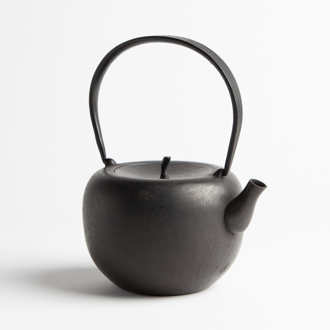 A black cast iron kettle on a white backround. The kettle is round with a flattish top and a lid a bit like the top of an apple. The kettle has a long handle for holding on top. 