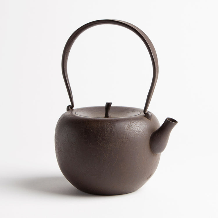A brown cast iron kettle on a white backround. The kettle is round with a flattish top and a lid a bit like the top of an apple. The kettle has a long handle for holding on top.