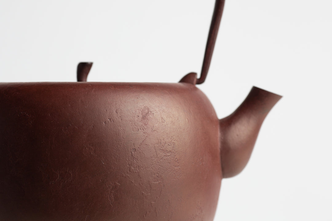 A close-up of a reddish-brown cast iron kettle on a white backround. The kettle is round with a flattish top and a lid a bit like the top of an apple. The texture of the cast iron is in focus.