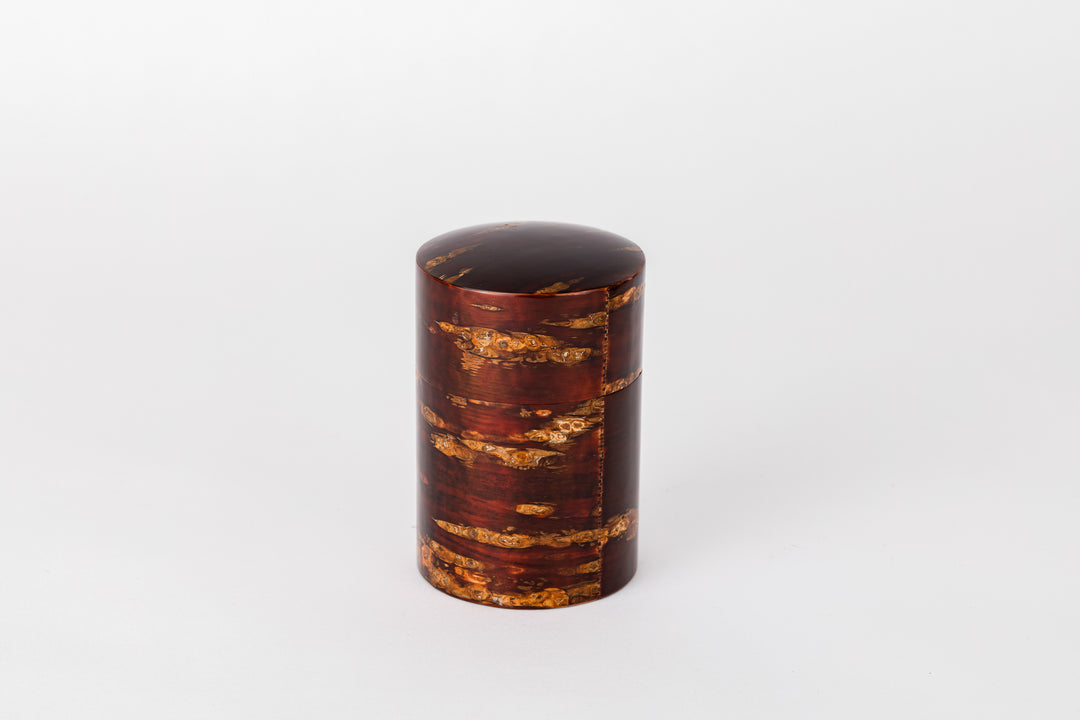 A cylindrical tea caddy made of cherry bark sits on a white background with its lid closed. The cherry bark has a natural red and orange striped pattern. It is polished and very shiny.