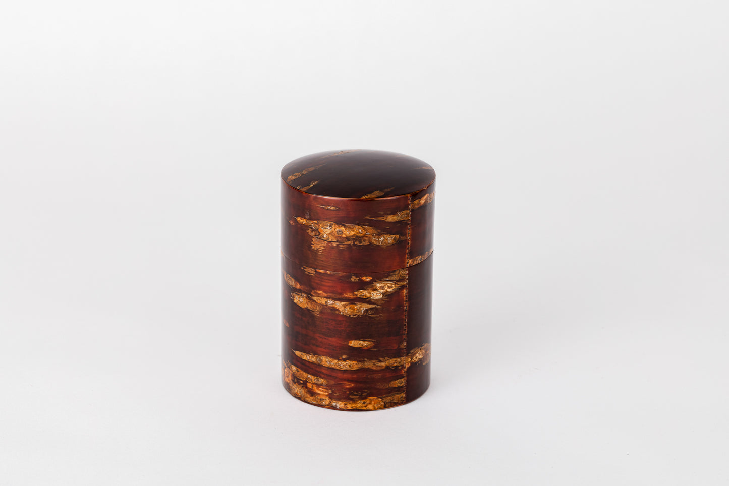 A cylindrical tea caddy made of cherry bark sits on a white background with its lid closed. The cherry bark has a natural red and orange striped pattern. It is polished and very shiny.