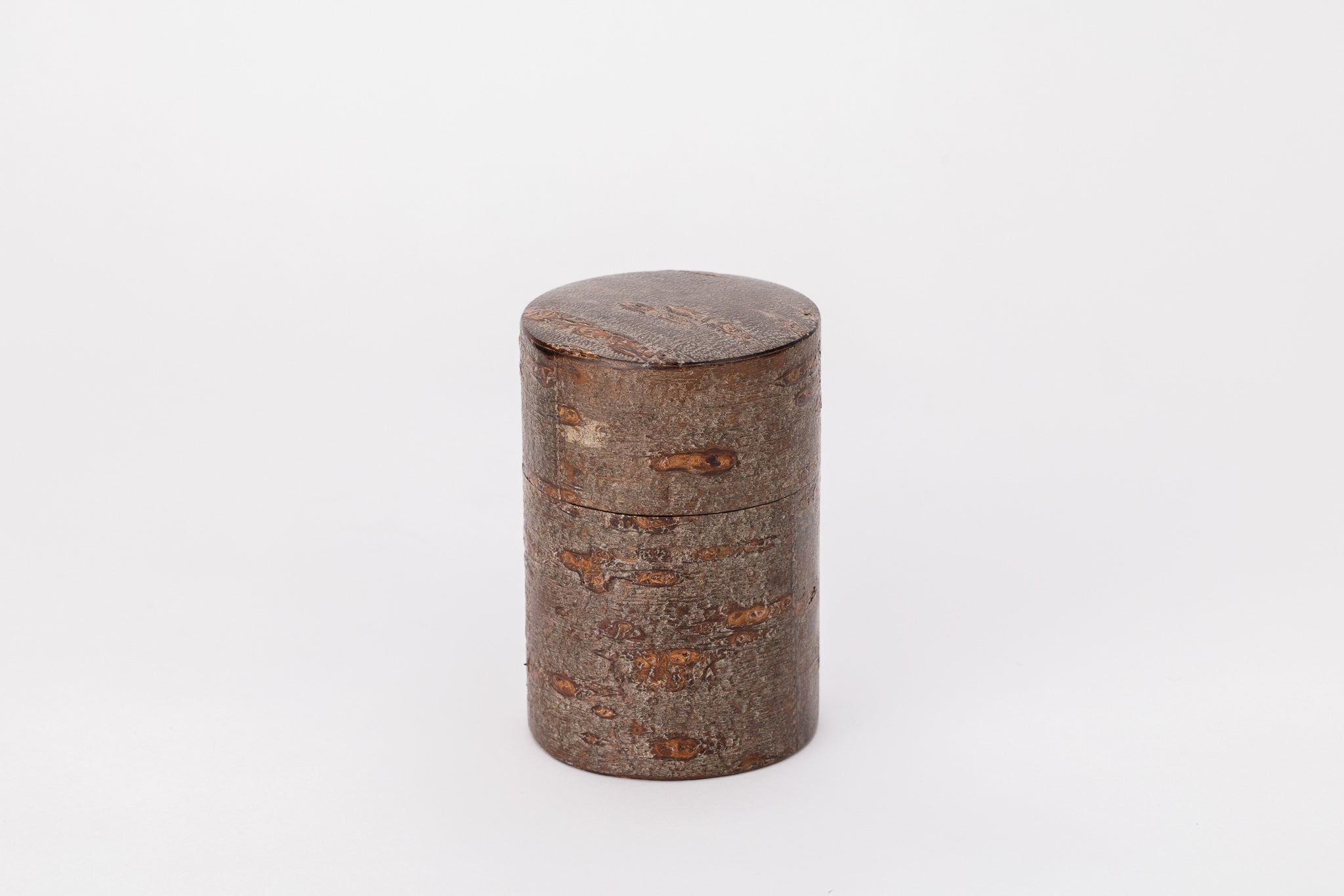 A cylindrical tea caddy made of cherry bark sits on a white background with its lid closed. The cherry bark is brown and rough like the bark of a tree.