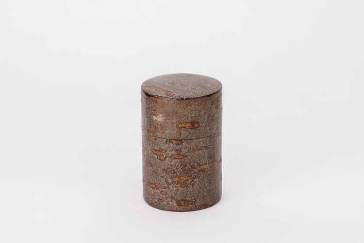 A cylindrical tea caddy made of cherry bark sits on a white background with its lid closed. The cherry bark is brown and rough like the bark of a tree.