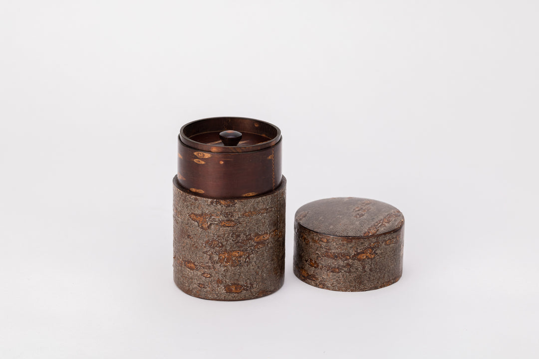 A cylindrical tea caddy made of cherry bark sits on a white background with its lid open. There is a second smaller lid sealing the inside of the cylinder. The outside surface of the cylinder is brown and rough like the bark of a cherry tree. The inside is smooth and polished and has a natural red and brown striped pattern. 