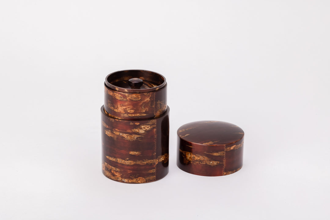 A cylindrical tea caddy made of cherry bark sits on a white background with its lid open. There is a second smaller lid sealing the inside of the cylinder. The cherry bark has a natural red and orange striped pattern. It is polished and very shiny.