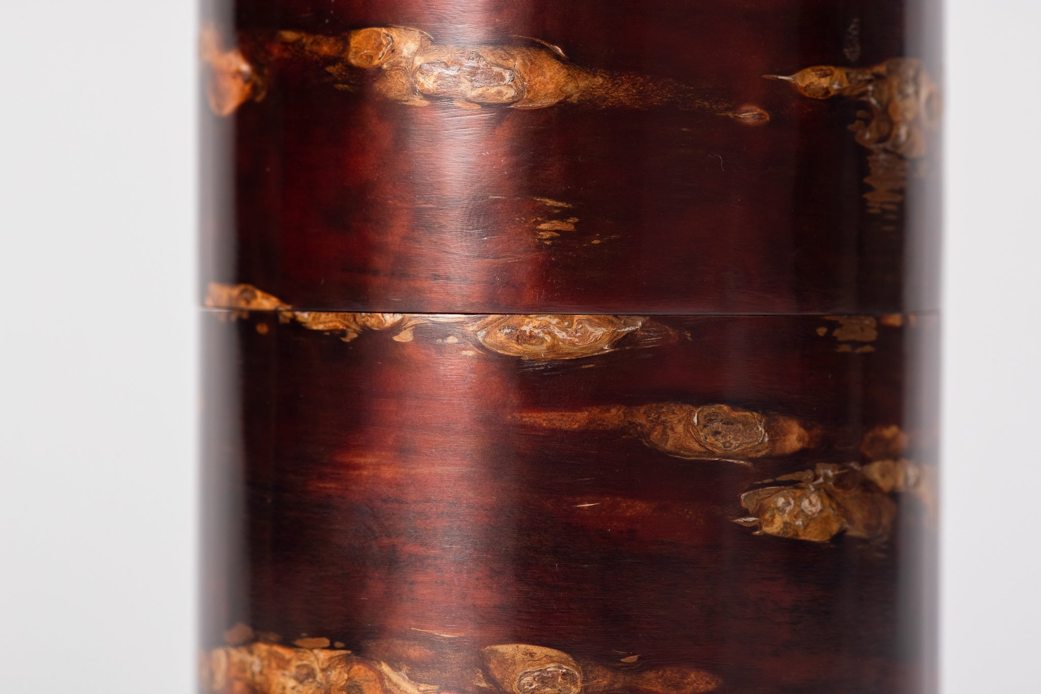 A close-up of a cylindrical tea caddy made of cherry bark sits on a white background.  The cherry bark is visible up-close and has a natural red and orange striped pattern. It is polished and very shiny.
