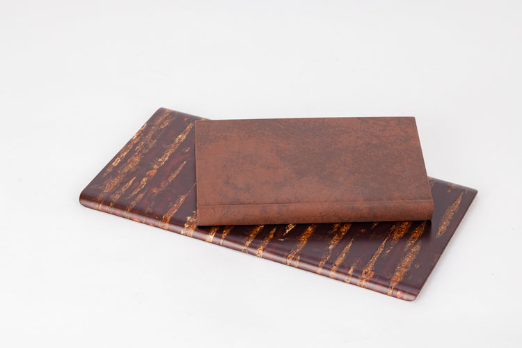 A rectangular wooden plate made of shiny red and orange cherry bark. A brown leather-bound book sits on top of the plate.