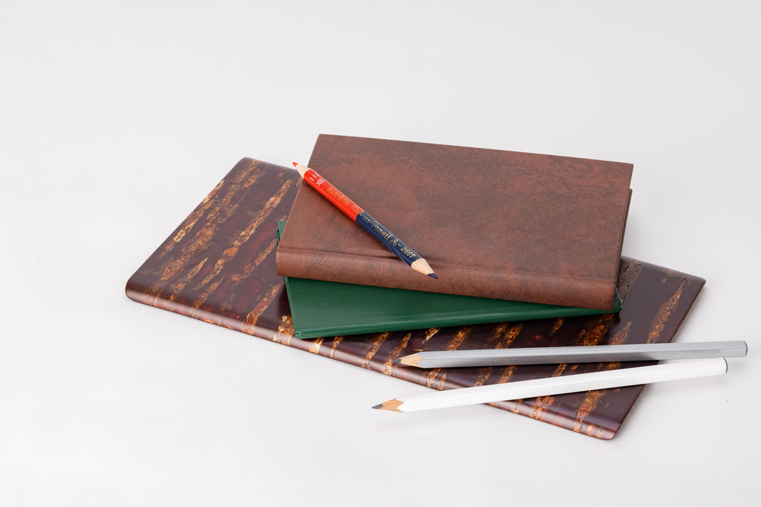 A rectangular wooden plate made of shiny red and orange cherry bark.  On the plate are a white and silver pencil, a thin, green leather-bound notebook, a thicker brown leather-bound notebook, and on on top of the brown book, a pencil crayon with one red end and one navy blue end. 
