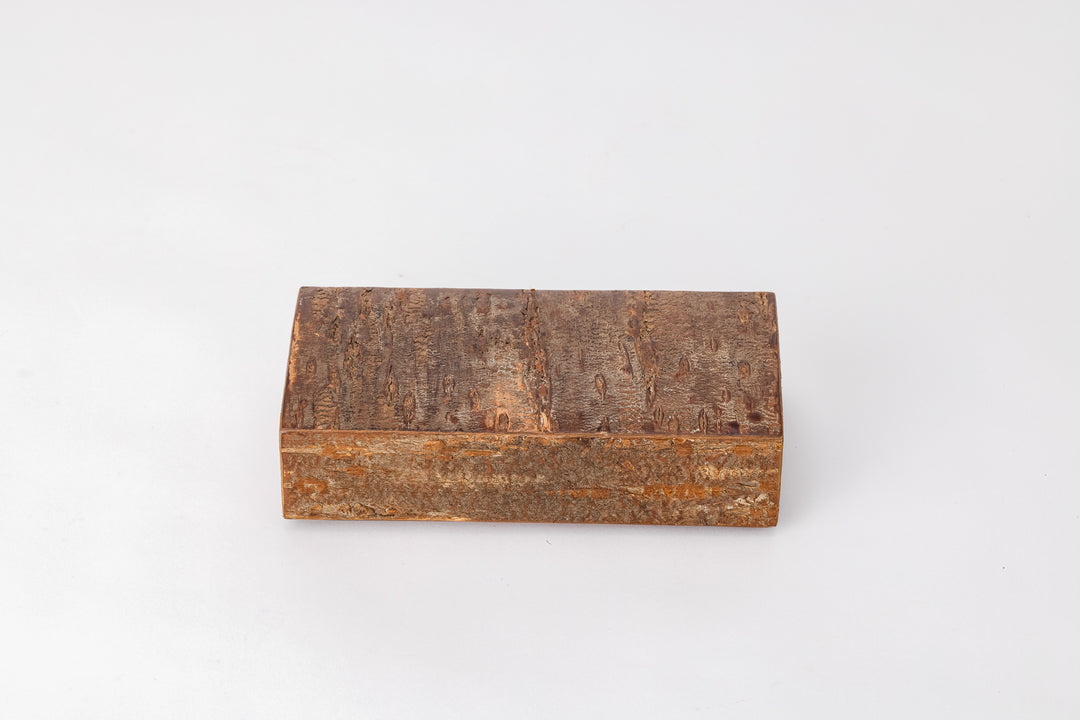 A rectangular wooden box made of cherry bark sits on a white background with its lid closed. The outside of the box is rough like the bark of a cherry tree. The long edge of the box is facing the vieewer.