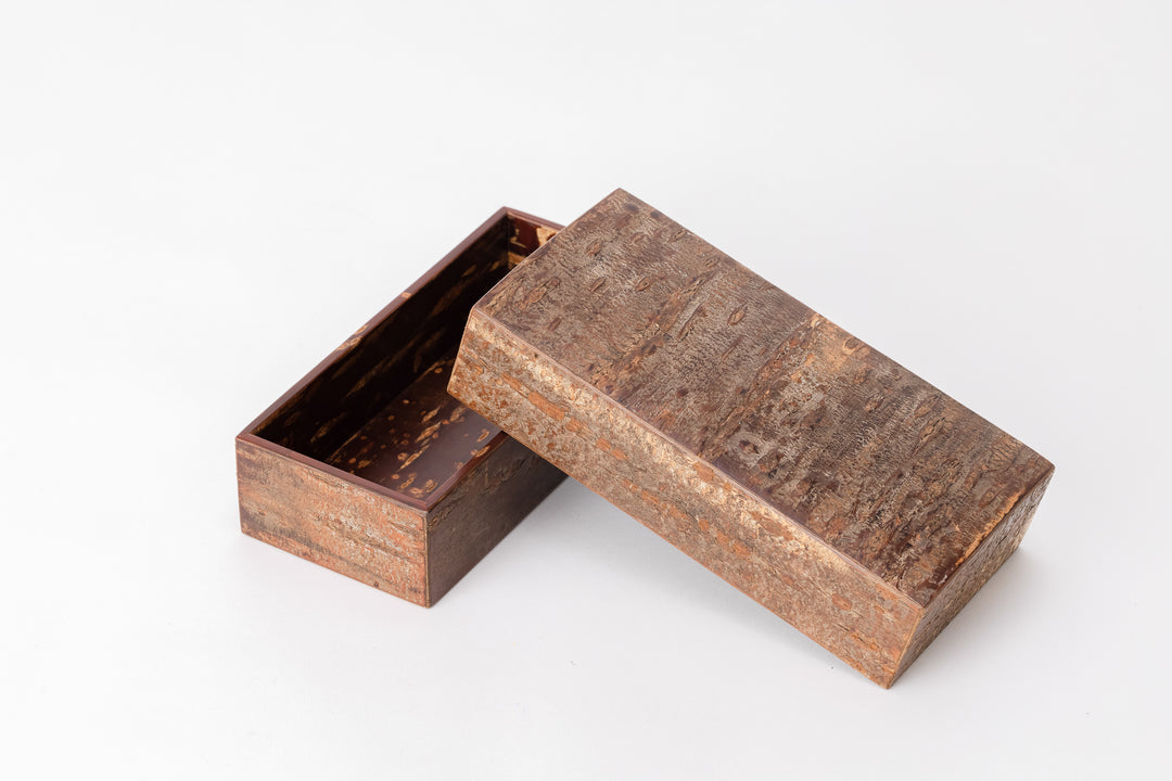 A rectangular wooden box made of cherry bark sits on a white background with its lid open. The outside of the box is rough like the bark of a cherry tree. The inside of the box is reddish-brown, polished and shiny. The box is placed diagonal to the viewer.