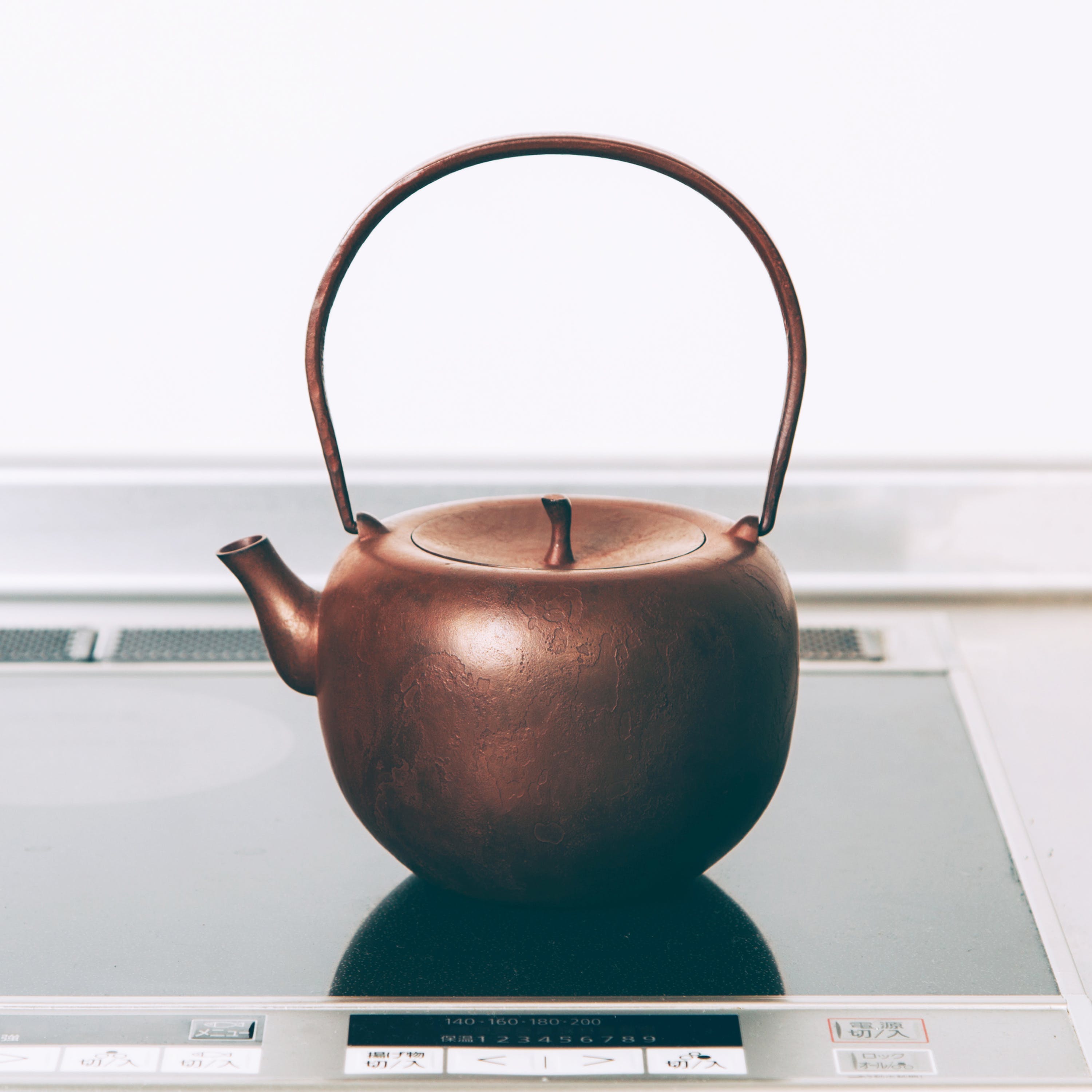 A reddish-brown cast iron kettle sits on a black induction stove. The kettle is round with a flattish top and a lid a bit like the top of an apple. The kettle has a long handle for holding on top.