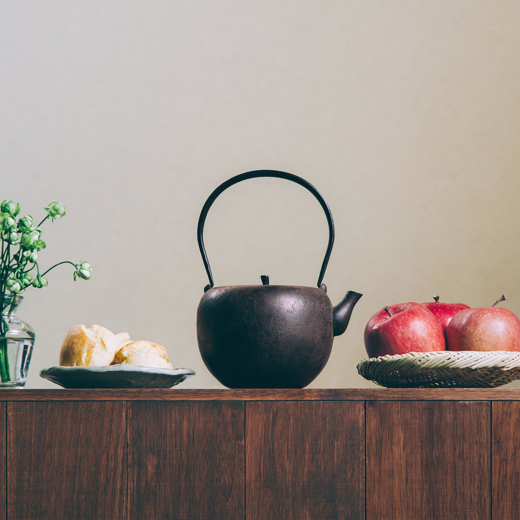 A wooden table is decorated with green flowers in a clear glass vase, a plate of scones, a basket of red apples, and in the center, a black cast iron kettle.  The kettle is round with a flat top and a lit a bit like the top of an apple. The kettle has a long handle on top.